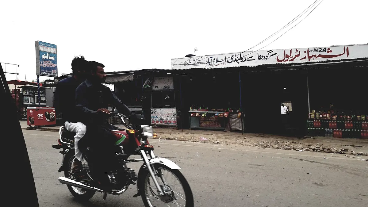 Two men riding a red 70cc motorcycle past roadside shops with Urdu signboards in Punjab